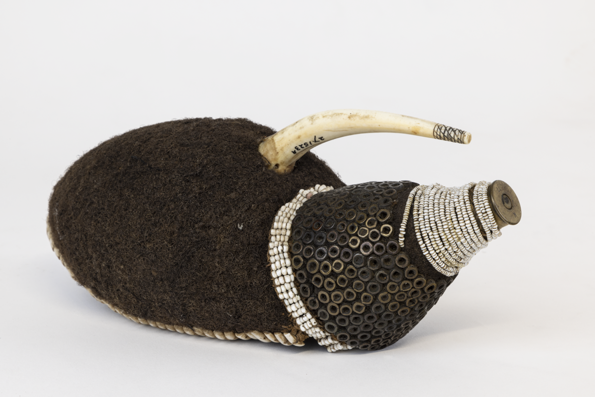 A hollow cap of felted human hair encircled by small pieces of cowrie shell threaded onto a single string. At the front is a curving ivory projection and cone with a foundation of human hair; the base is covered in four rows of annular white beads.