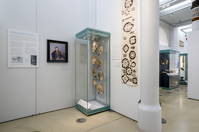 Installation view of the exhibition with barkcloth and fans visible next to a portrait of von Hügel.