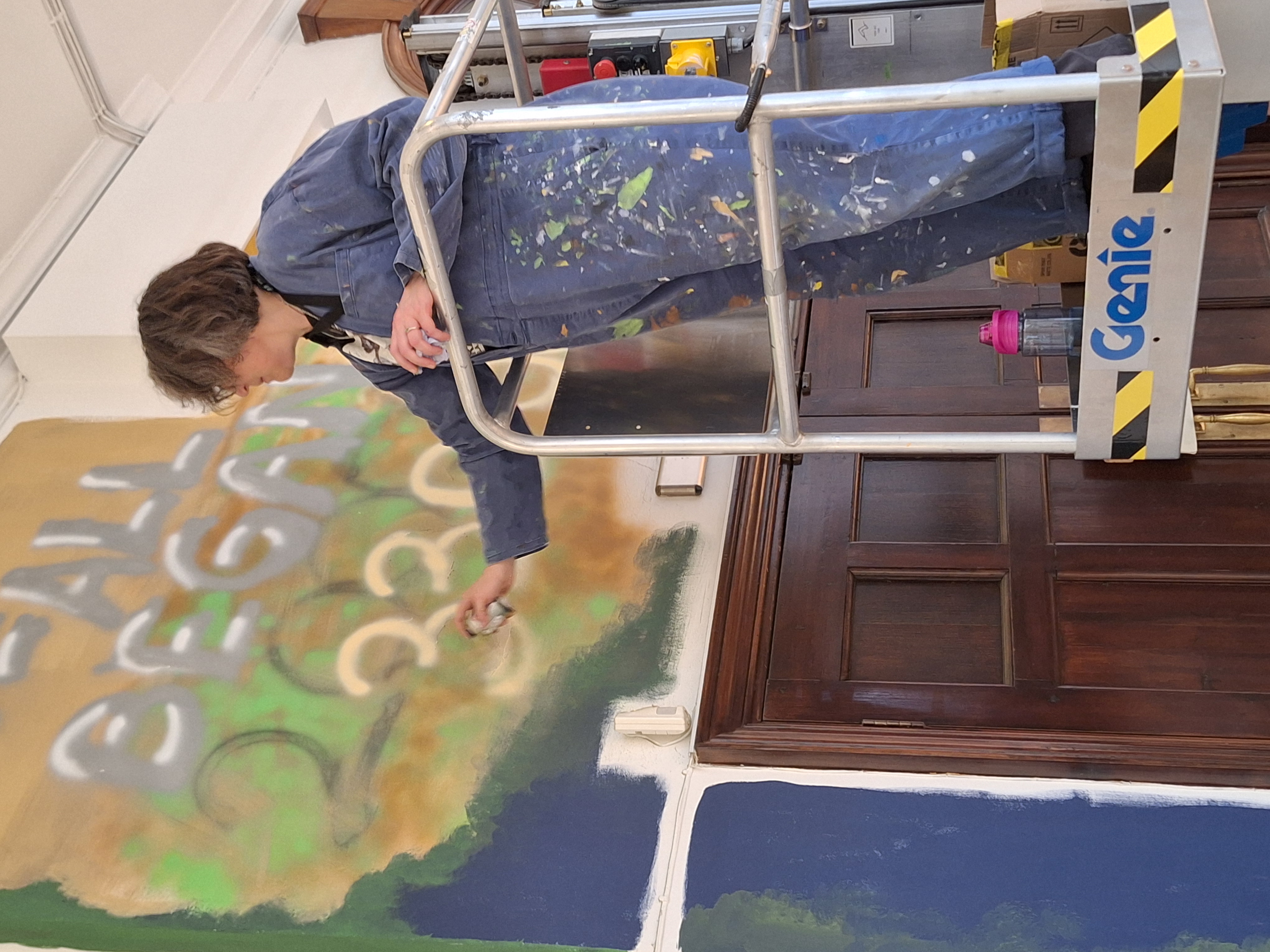 An artist stands on a hydraulic platform as she adds to a graffiti mural above a pair of closed wooden doors.