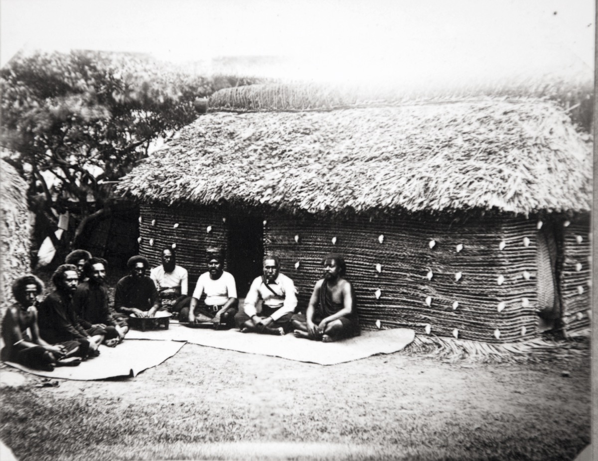 Black and white photograph of nine men sitting cross legged on mats outside a house decorated with shells.