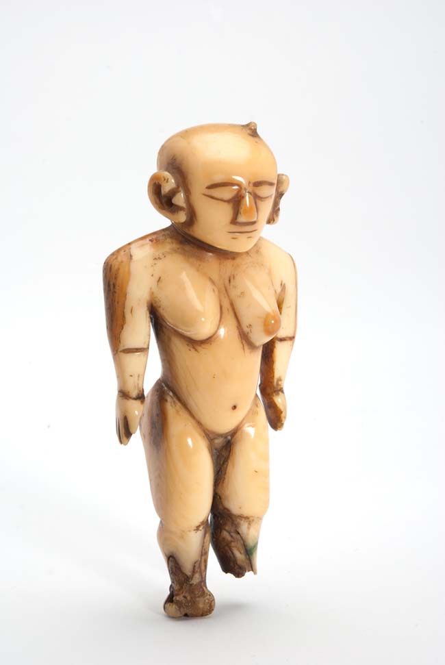 Small female figure carved from a single piece of whale ivory. Standing, straight legs and arms along the body. One foot missing, the other damaged.