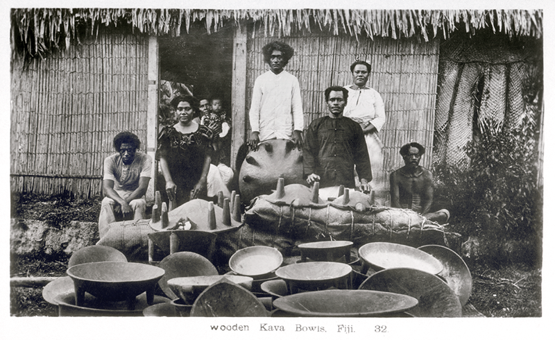 Black and white photograph of men and women standing in front of large bowls, in front of a reed-thatched house.