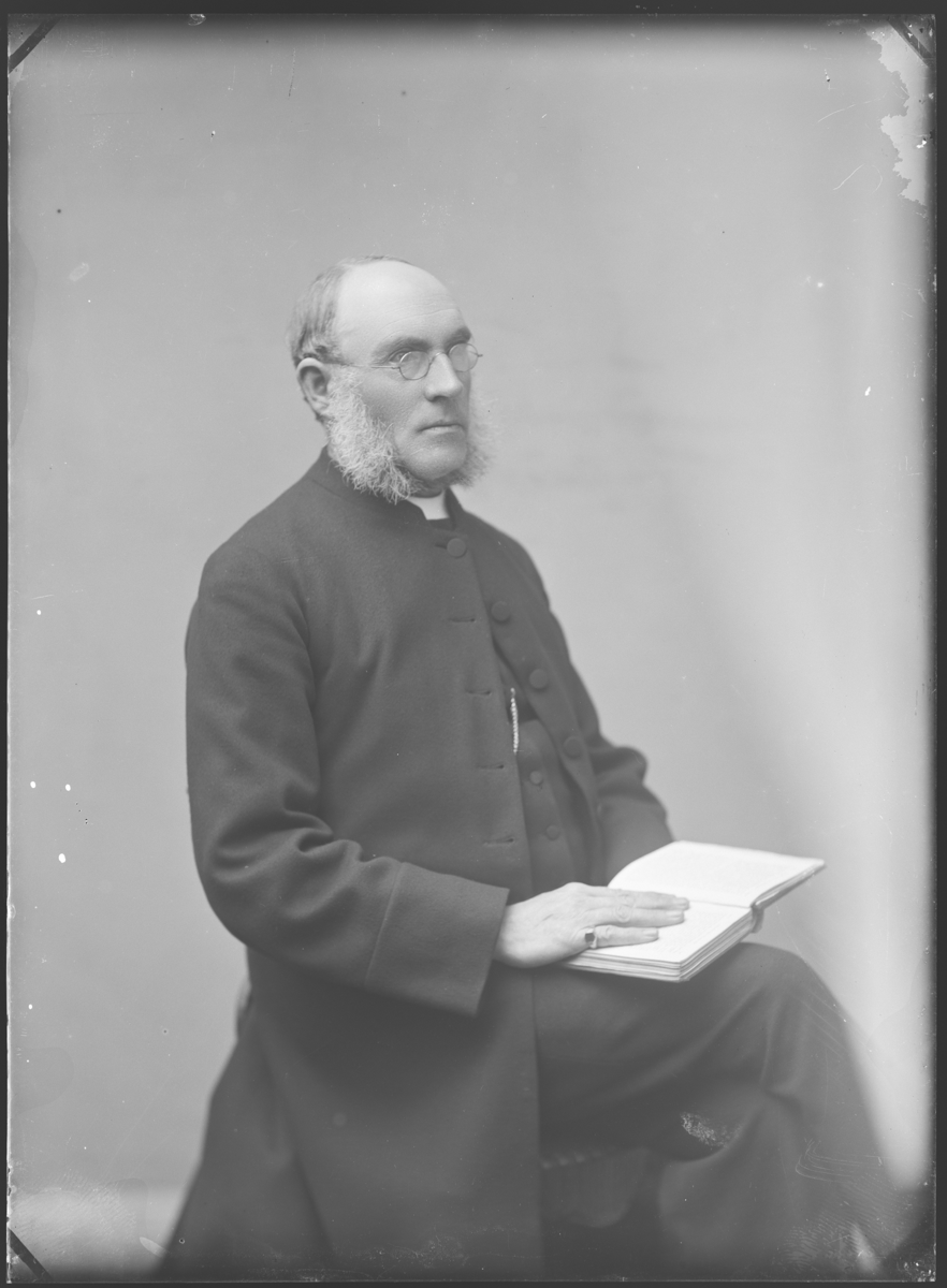 Portrait of a priest, seated, holding a bible. Black and white negative. He has a white beard and wears glasses, as well as a signet ring on his right hand.