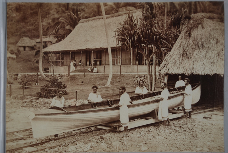Sepia photograph of men wearing white garments pushing a boat across tracks, out of a thatched roofed building. A large house can be seen in the background.