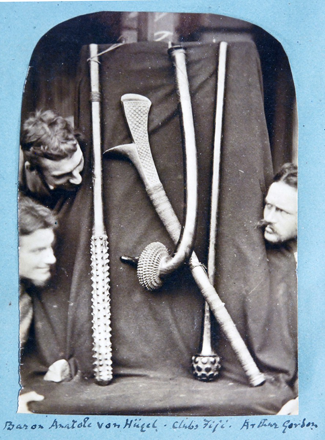 Three men peer round a cloth-backed display of clubs and other items. Photographed backed against blue paper