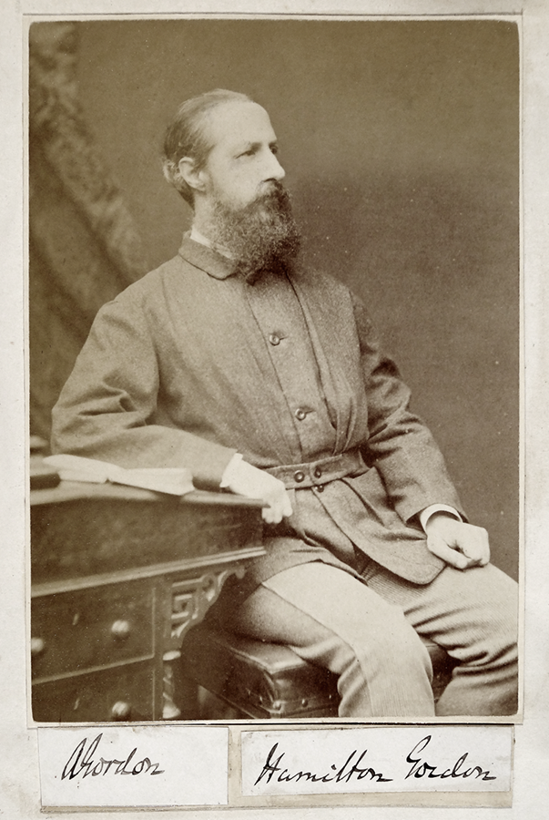Sepia photograph of a white man with a large beard dressed in victorian-style clothing, looking off to one side, seated next to a dresser. Labels saying 'Gordon' and 'Hamilton Gordon' are seen beneath the photo.