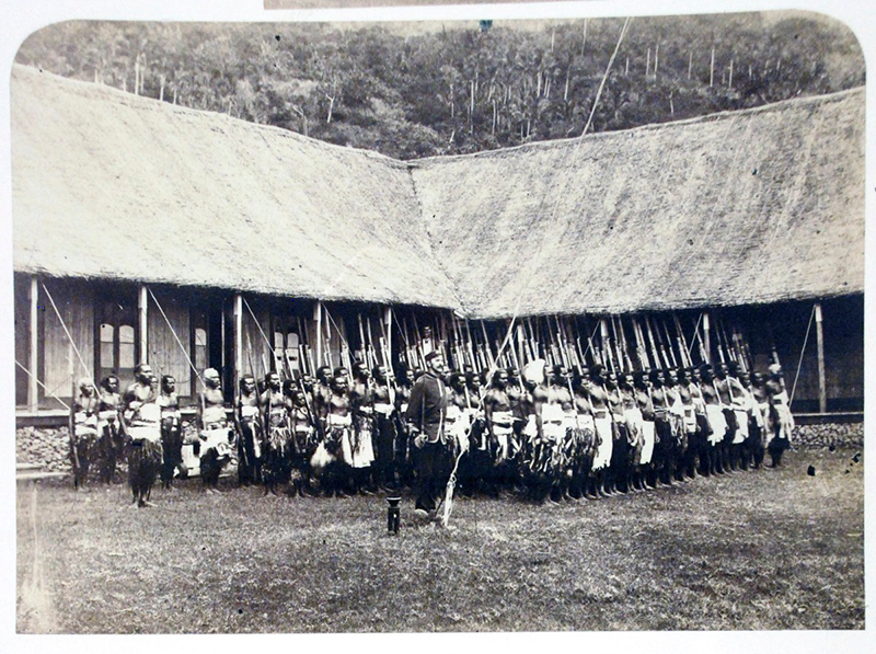 Black and white photograph of a group of people standing in line, with long spears, in front of a house