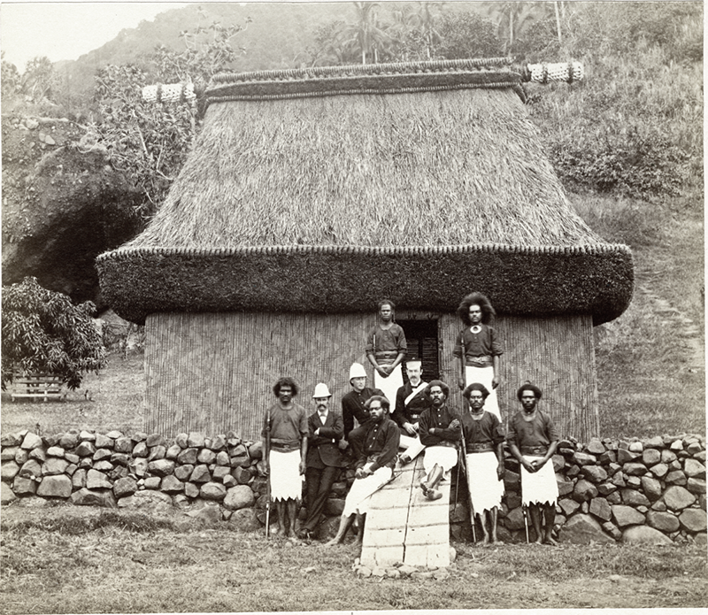 Photograph of a group of men sitting and standing in front of a thatched roof house