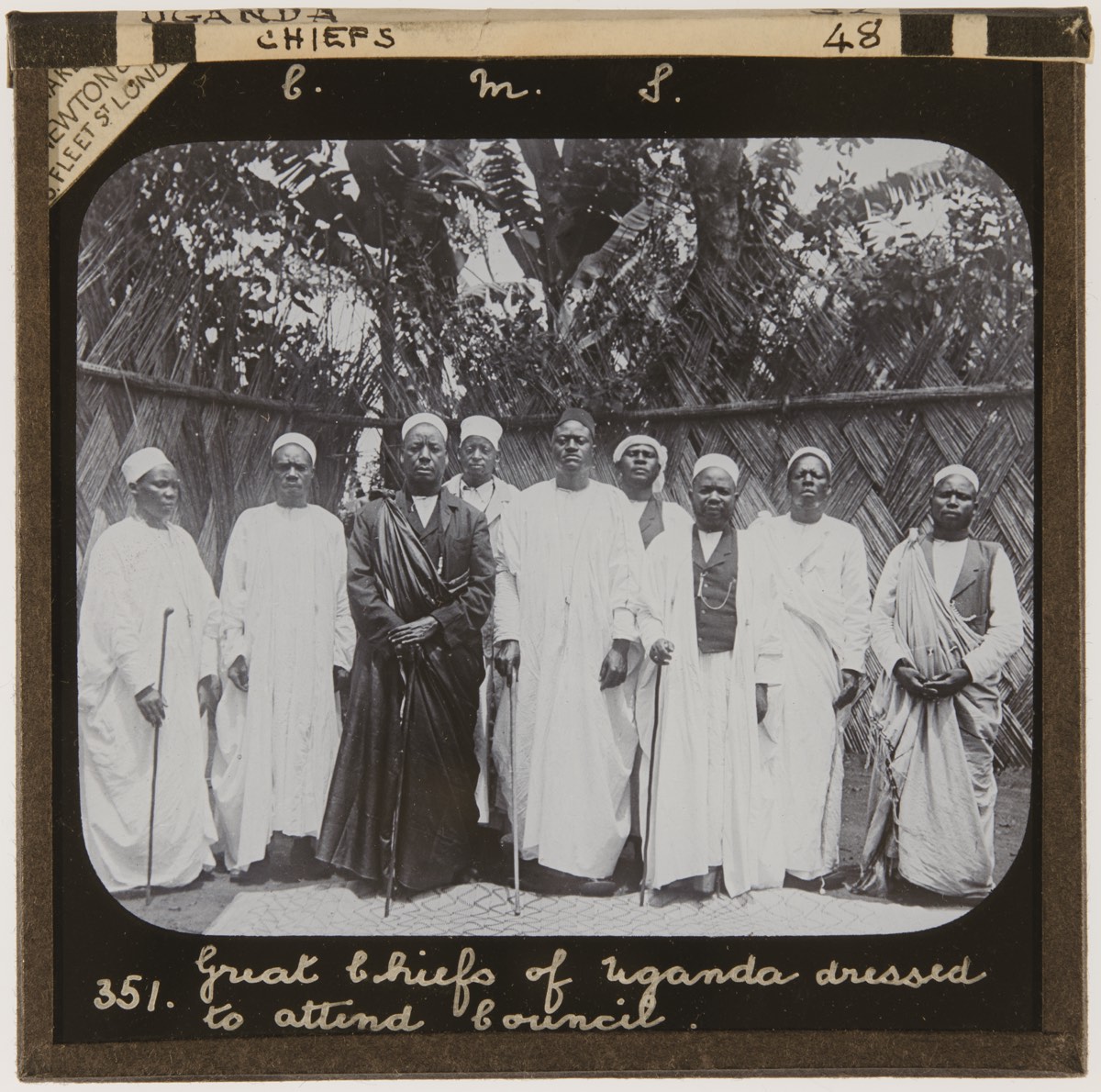 Black and white group portrait of dignitaries and chiefs, mostly in white clothing and one in black. Photograph mounted on a photo slide, with 'Great chiefs of Uganda dressed to attend council. 351' handwritten at the base.