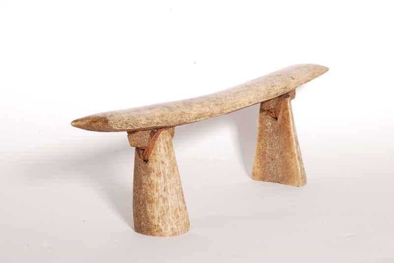 Headrest made of whale bone, with a rounded top and the ends slightly curving upwards. Two legs of the same bone are attached by lugs at the back.