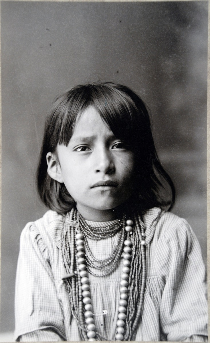 A studio photograph of a Navajo child, with shoulder length dark hair and wearing many necklaces over the top of a checked cotton blouse or dress (uniform).
