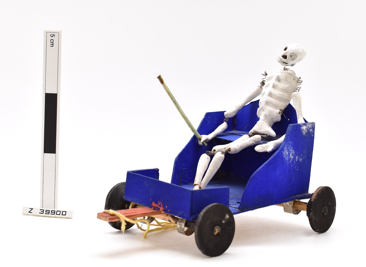  Model of a skeleton sitting in a blue cart. The skeletons joints are made from springs and it is holding a stick. 