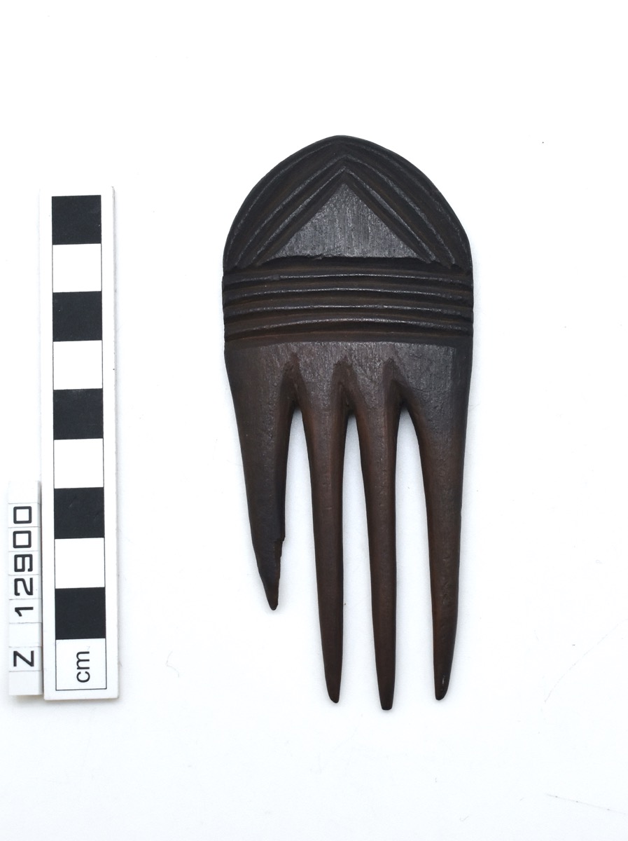 A comb carved from wood with four cylindrical and tapering prongs. The handle has a rectangular section then a constriction from which it flares outward and a triangular shaped terminal. One side is decorated with carved lines.