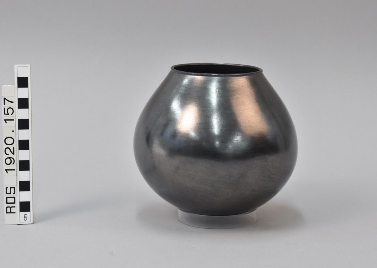 Highly glazed ceramic milk pot, silver in colour, photographed against a studio background. A measure shows it to be around 15cm in height, and shows the accession number 'ROS 1920.157'.
