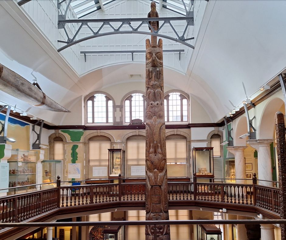 The Andrews Gallery with cases displaying world archaeology collections. The gallery has a balustrade around the space in the centre of the room, which opens out into the gallery below. At the far end of the gallery a totem pole rises from the floor below