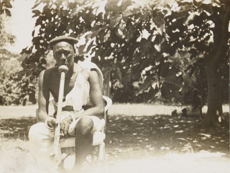 Asepia photograph of an old man, Reth Fatifi Wad Yor, smoking a pipe. He is sat on a chair in front of a large sun-dapped tree scene.
