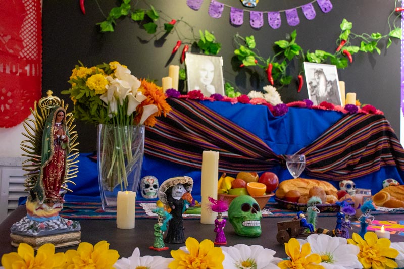 The middle and top tier of a Día de Muertos (Day of the Dead) altar. It is decorated with flowers, food, skeletons, skulls, a religious statue (possibly the Virgin Mary) and a couple of black and white photos of the deceased whom the altar honours.