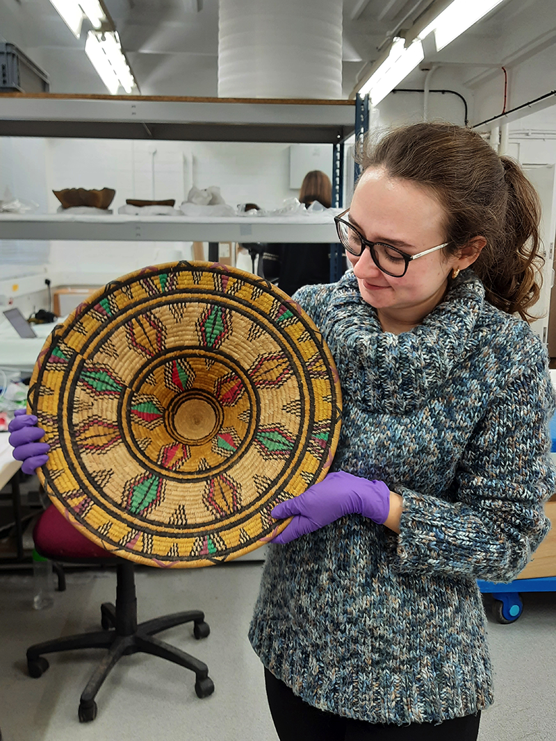 Emily Shorter, holding a large colourful woven basket. She is smiling at the object, and wears a blue knitted jumper, and has purple conservation gloves on. She stands in the MAA workroom.