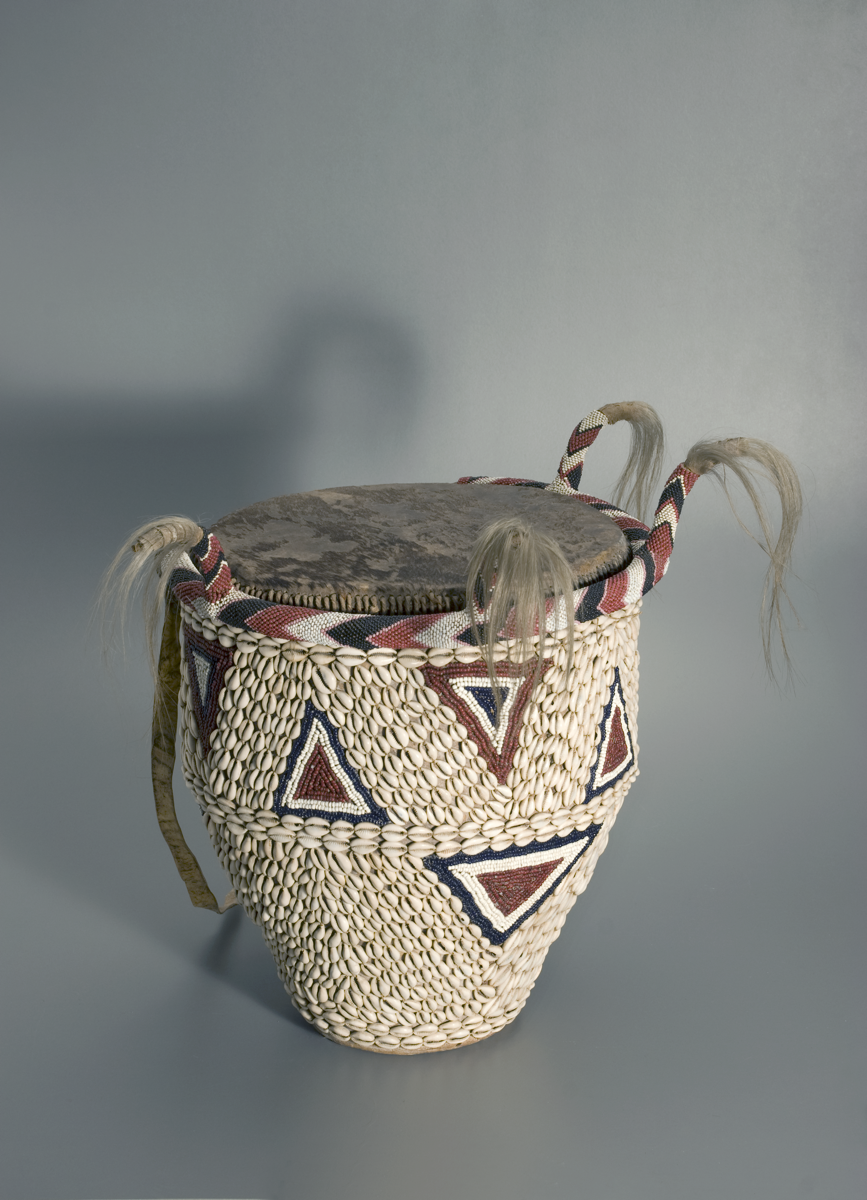 Royal drum, decorated with cowries and triangular designs in red and black glass beads. Around the top is a beaded band with four beaded vertical projections, terminating in hair tufts.