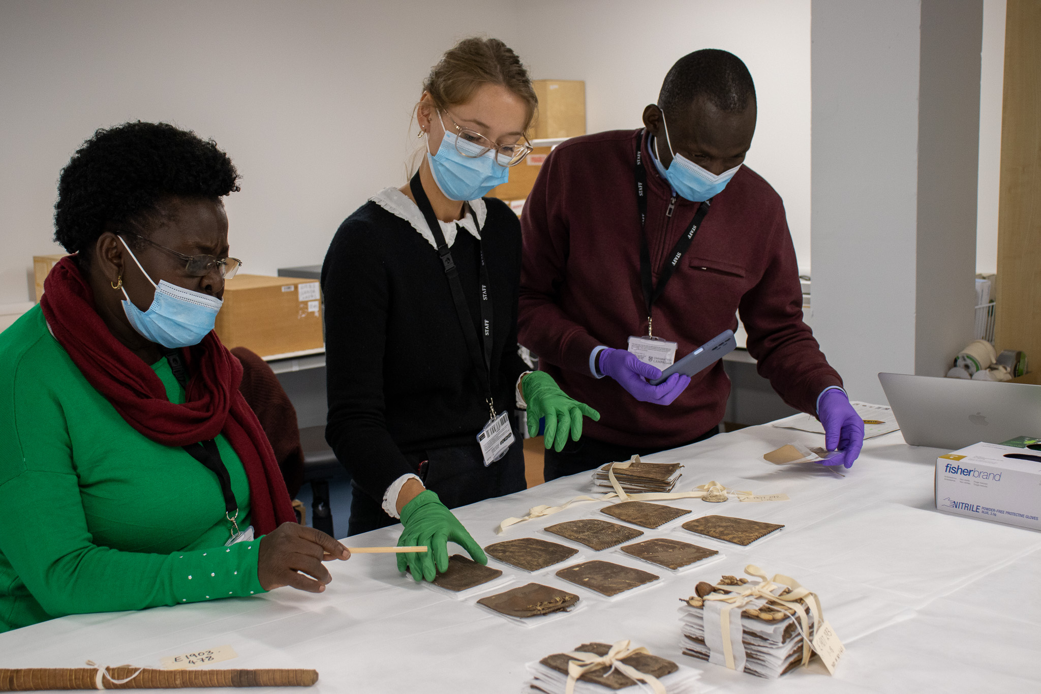 The former Commissioner for Museums and Monuments and Principal Curator, Uganda Museum, in the museum's off-site store looking at a set of leather cards. Between them stands a Collections Assistant. All three wear face masks.