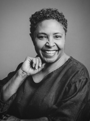 A widely smiling curator looks towards the camera. The image is black and white and she has short dark hair and wears a dark top. Her right handis raised and rests near her jaw.