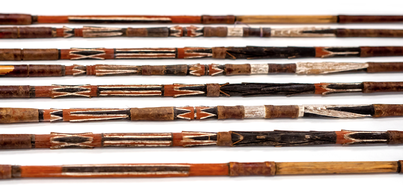 The wooden shafts of a collection of arrows which have been carved and painted using red, white, and black pigments.