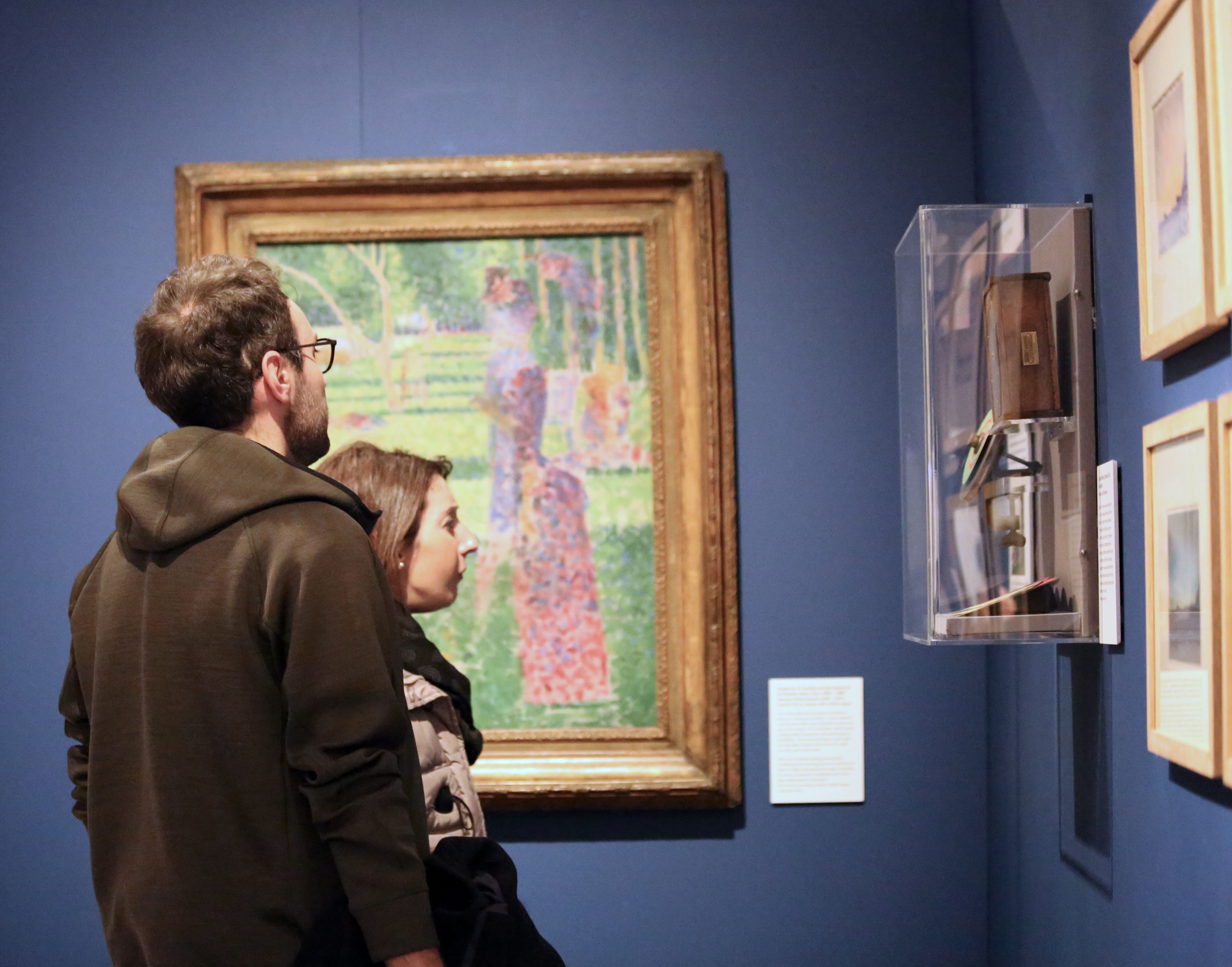 Two visitors to the museum stand in front of a painting. They are looking at objects displayed on the wall adjacent.