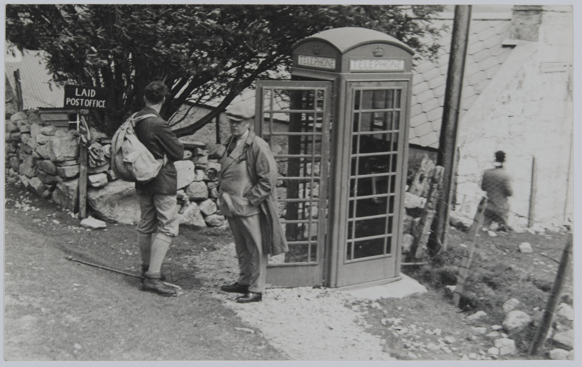 Black and white photo of James Wordie and a second expedition member standing next a British telephone box and a sign pointing to “Laid Post Office”. Village buildings are in the background.