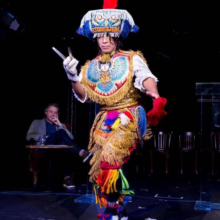 A dancer performs, possibly on a stage. They wear a brightly coloured Peruvian costume and hold a pair of scissors in one hand. There is a large rectangular hat on their head depicting the face of a bird, possibly an owl. There is a full depiction of the 