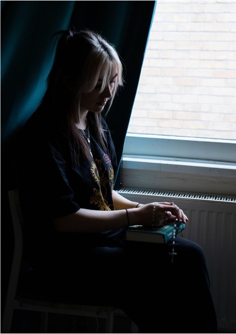 Photograph of a woman sat down, holding her grandfather's possessions, against a window. The lighting is deep and melancholy.