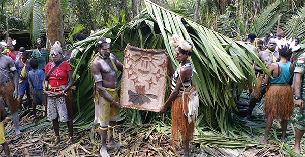Asmat villagers hold a pit mat in the forest. A man and a woman hold the mat with people surrounding.