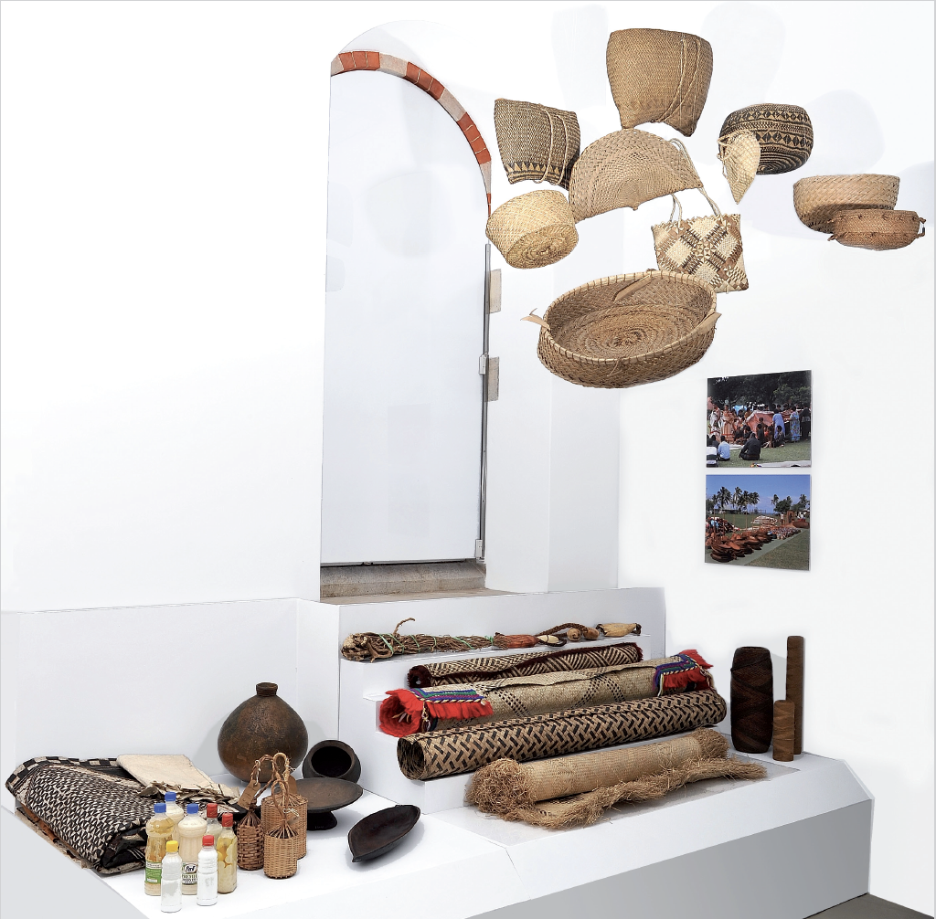 Installation image. Baskets hang from the ceiling, photographs are on the wall, barkcloths lay on the base alongside other objects of interest.