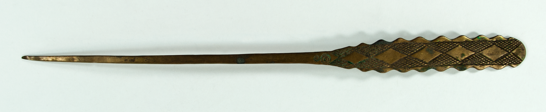 Brass hairpin with serrated style edges and a rounded end. Decorated with vertical lines and diamond shapes surrounded by crosshatching. The shaft of the pin is long and narrow.