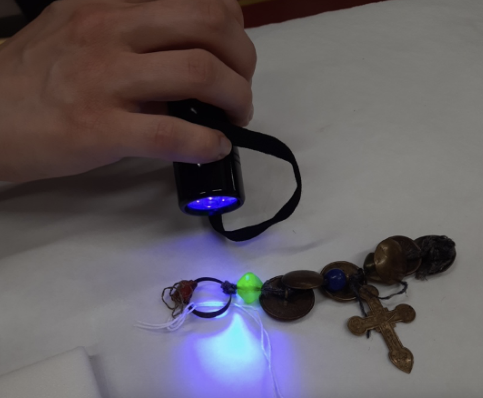 A hand holding a torch shines UV light onto a necklace revealing a bead which glows green.