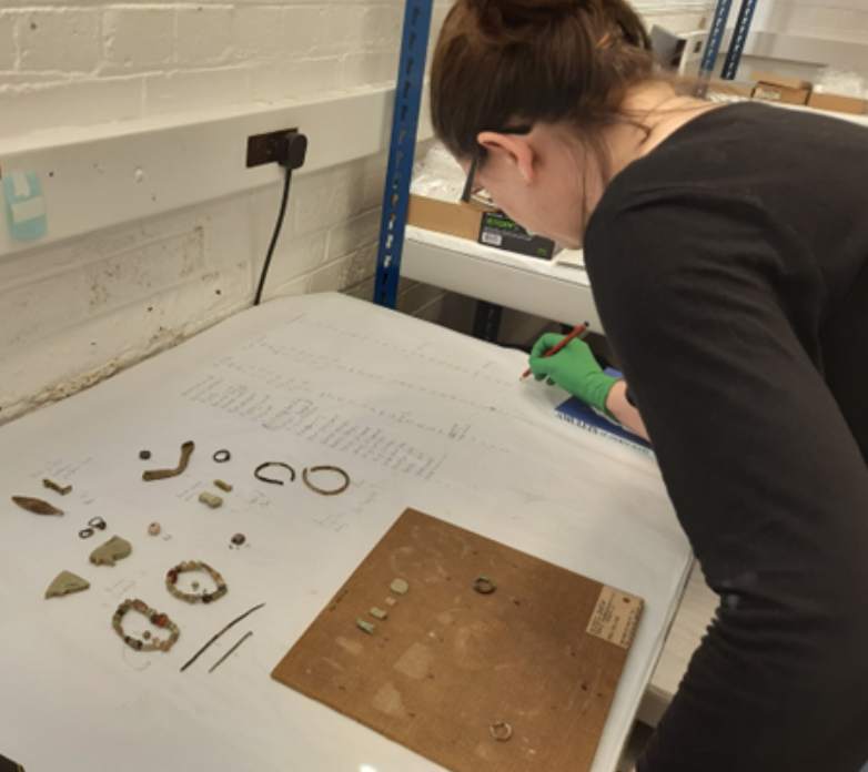 A member of staff works on an old museum display board. Some of the objects removed from the board lie in front of her as she works on a list to identify them all.