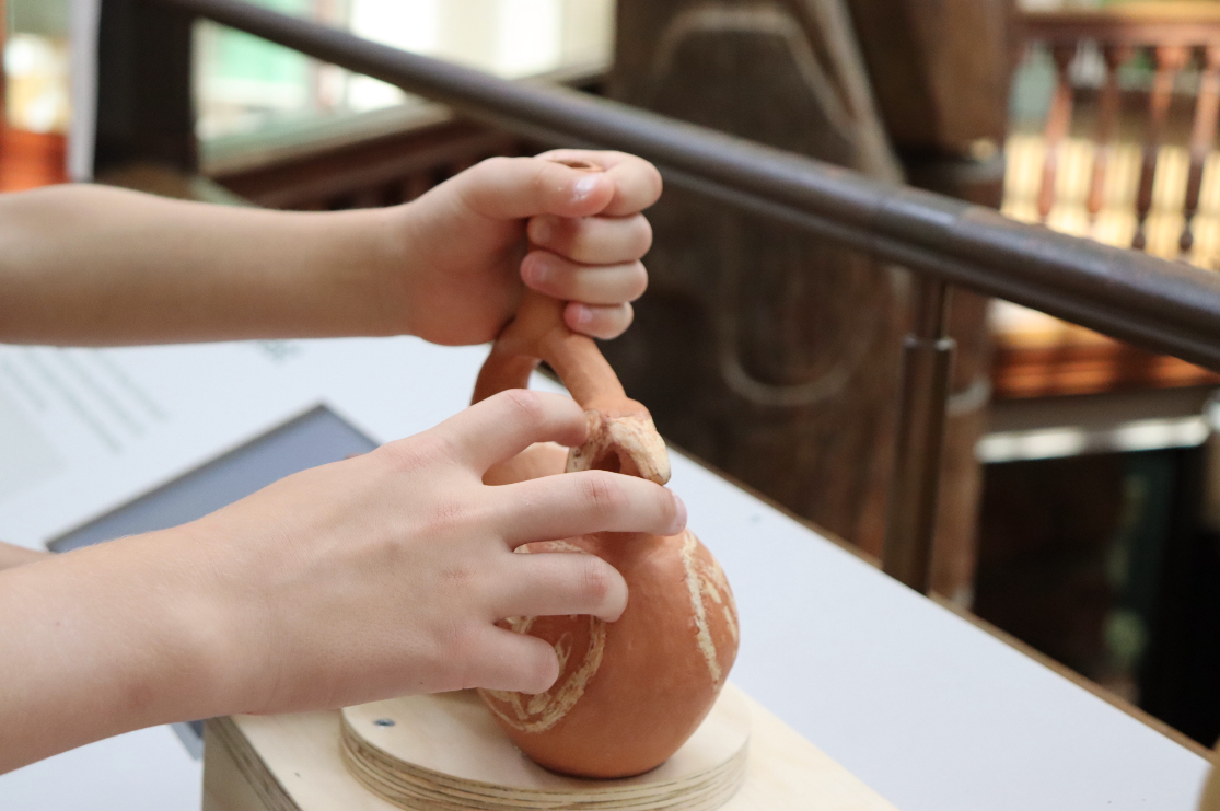 A child's hands touch a replica of a terracotta vessel consisting of two globular chambers with a spout in the centre and a moulded parrot's head at one end.