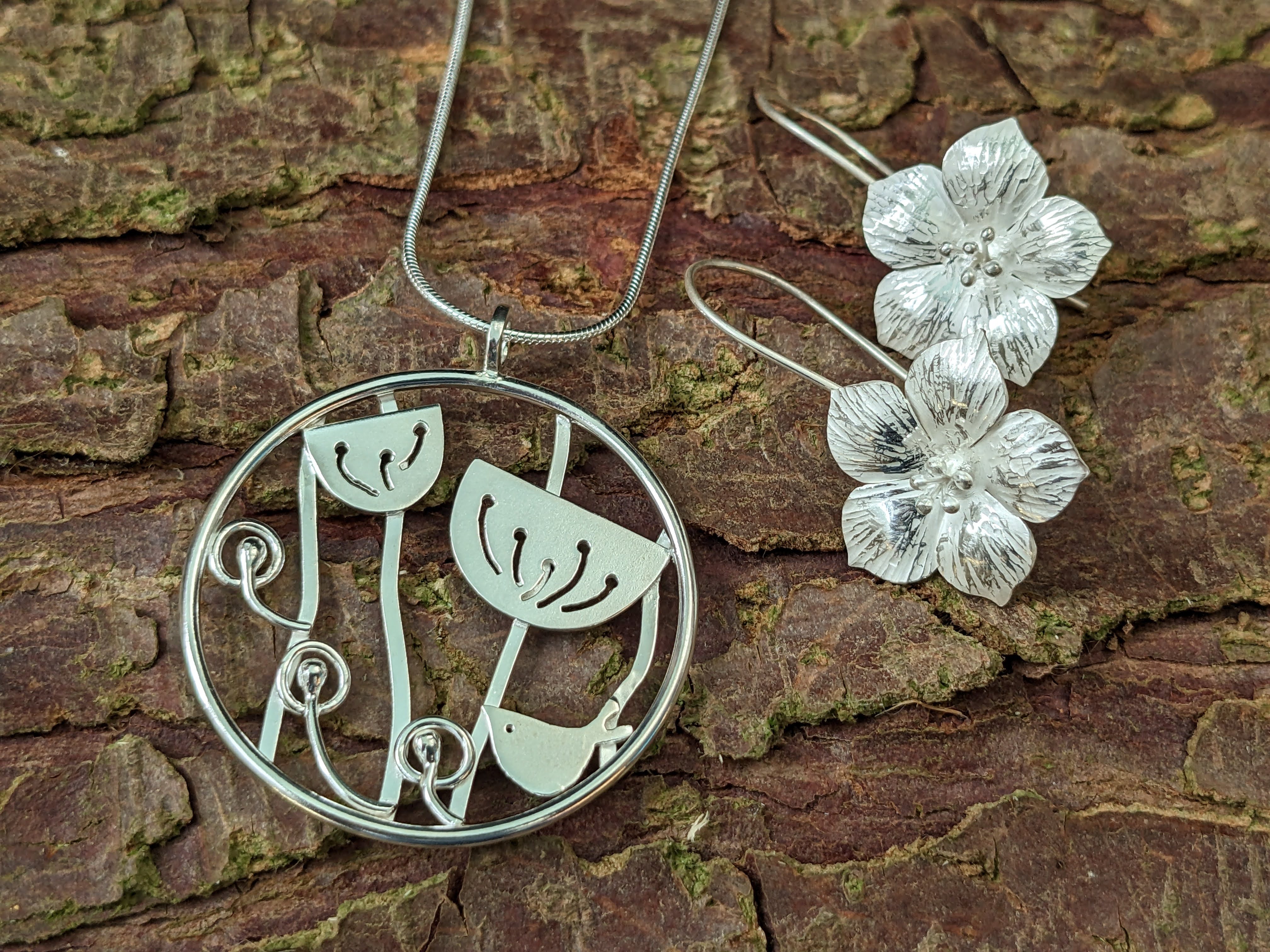 A silver pendant depicting a bird and seed heads and a pair of flower-shaped earrings rest against tree bark.