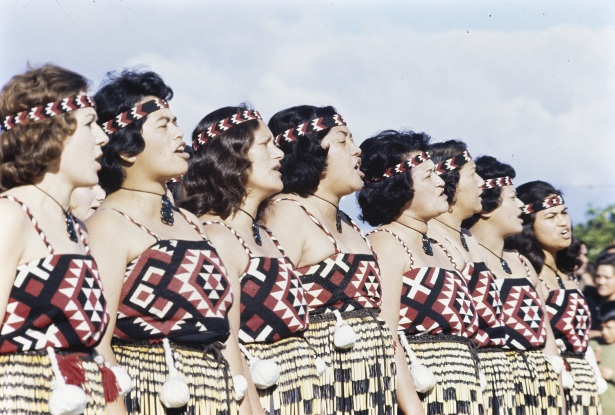 A group of 8 Māori women performing a dance. They are wearing tops, and headbands in black, red and white patterns, piupiu (skirts), and hei tiki (pendants). They are standing shoulder to shoulder with hands by their sides and appear to be vocalising.