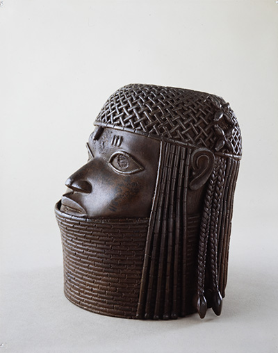 Finely cast bronze head with a plaited headdress and neck rings up to the bottom lip and extending far down. The hair is plaited and hangs down to the sides of the head. Object is viewed side-on.