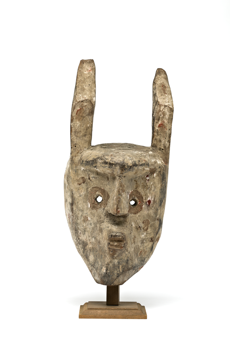  Carved wooden mask, painted white and red, with two long ears or horns and eye holes. The face has both animal and human features, with a small mouth with bared teeth, a straight nose, and two circular eye holes either side.