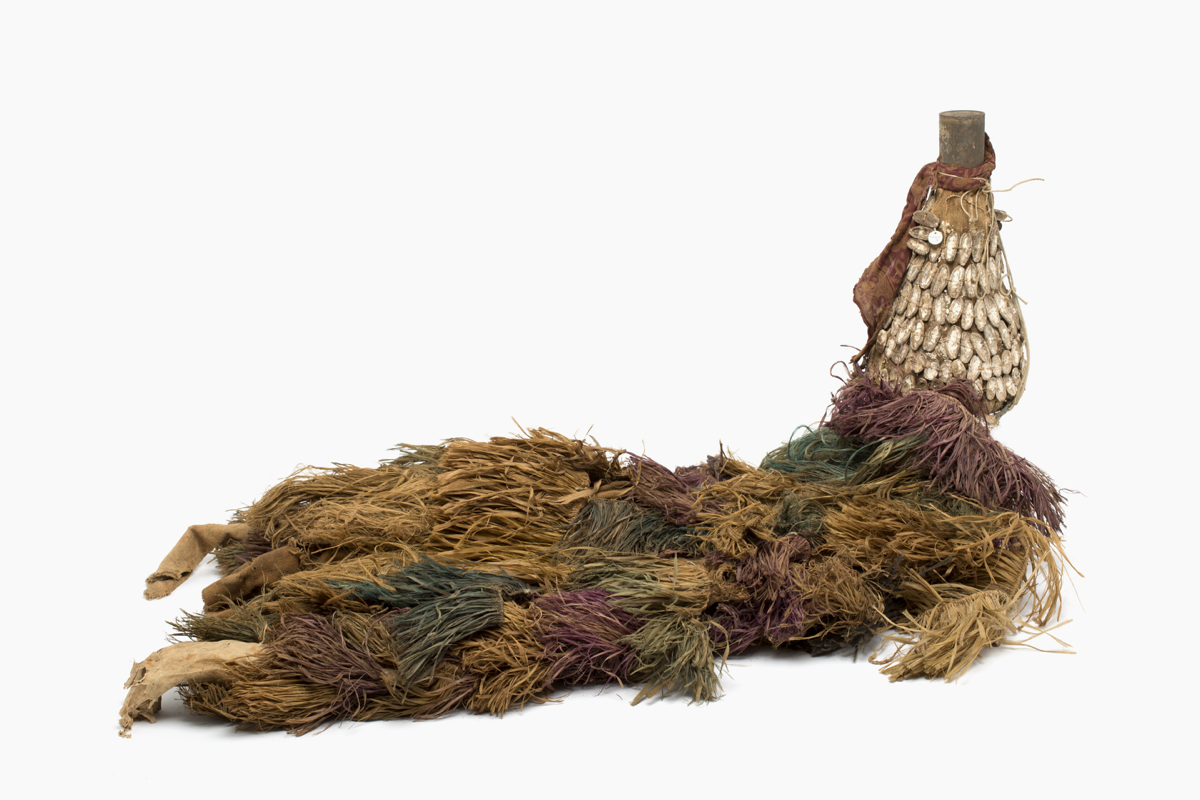 Masquerade costume made out of raffia and cloth. There is a basketry conic helmet covered with cloth and half parts of nut shells. The top of the hat has a rusty can. The colours are green and brown.