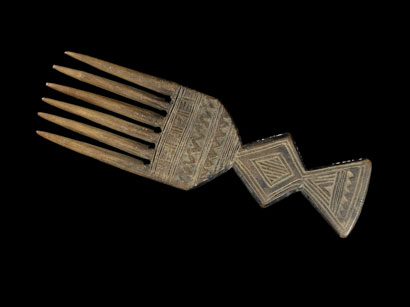 Wooden comb with six prongs, against a black background. Both sides of handle and shoulder carved with different pattern.