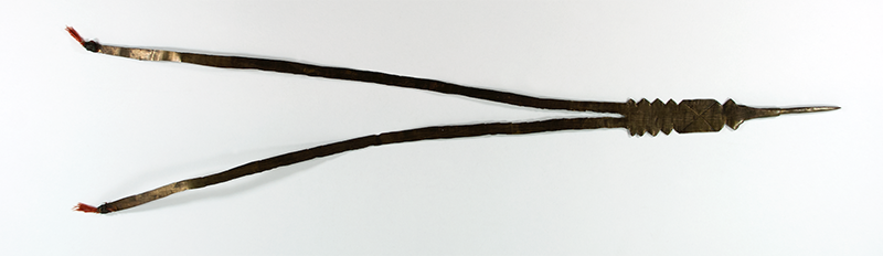 Brass hair ornament for dances. The sheet of metal splits into two long tails, each clasping a red feather.