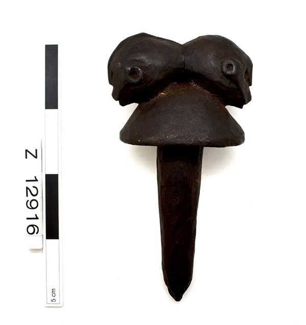 Bottle stopper, with a rounded top and two back-to-back heads above. Stopper has been smoothened by hand through use. Dark brown.