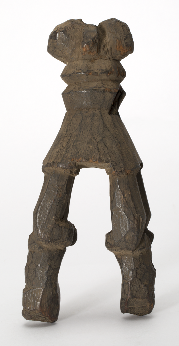 Loom attachment carved from wood. Abstract carving possibly depicting two human figures arranged back to back, features indistinct. Bottom section diverges and each side is carved through to appear like pairs of legs. 