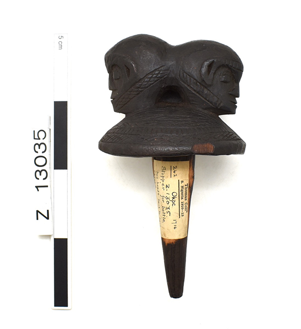 Bottle stopper, rounded top with two back-to-back heads. Features incised designs along foreheads and jaw lines. Chip in rim of top. Dark brown.