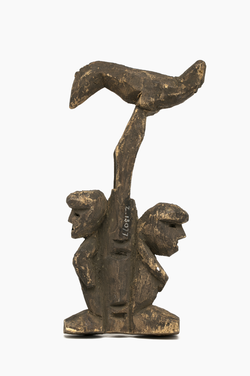 Wooden carving of two figures standing back-to-back at the base of a post or tree with a bird at the top. One of the figures has its hands across the front of the body, while the other has the arms by its sides.