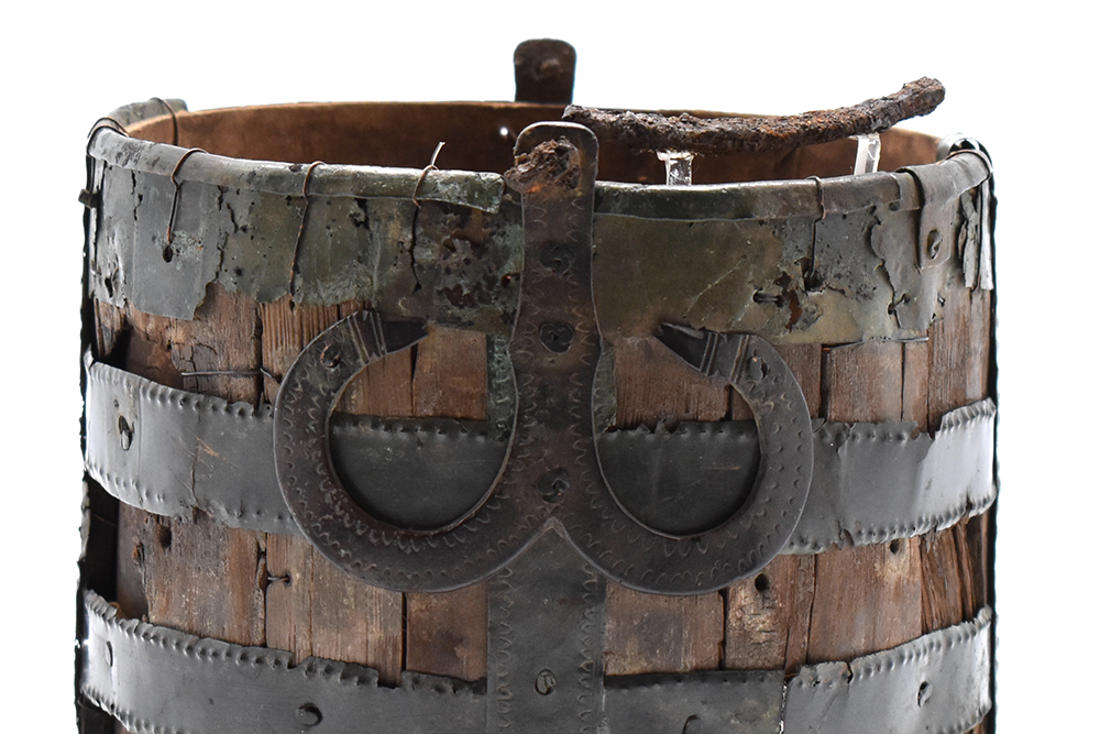 Cropped or close up image of the bucket, showing the weathered wood and hammered details of the metal bracing, with a double horseshoe shaped loop figure at the centre front.