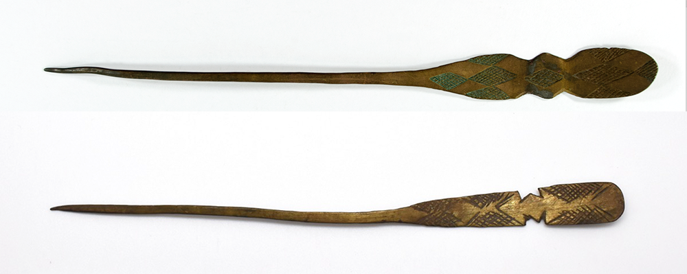 Two brass hairpins. One is rounded at the end and decorated with crosshatching, the other is a brighter gold/brass and the head is rounded at the end with an 'M' shaped notch at each side.