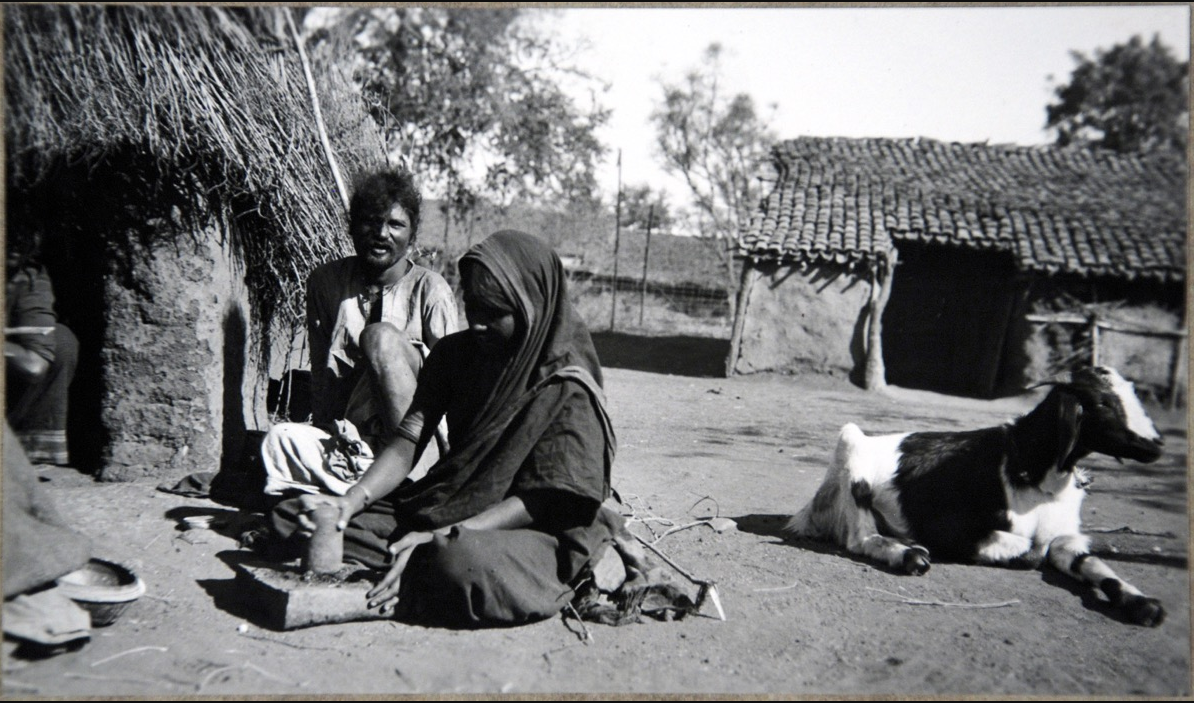 A woman wearing a sari uses a pestle and mortar to grind spices. Behind her sits a man whilst a goat lies off to the right.
